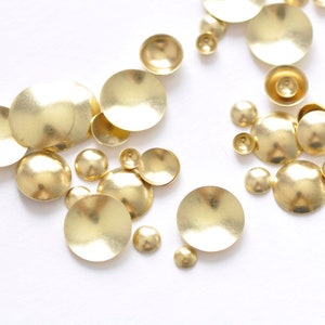 Op de afbeelding: Een assortiment kleine, goudkleurige metalen versieringen in verschillende maten, verspreid op een wit oppervlak. De vormen omvatten platte schijven en afgeronde, koepelvormige vormen, die geschikt zijn voor knutsel- of decoratieve doeleinden.