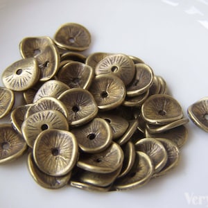 以下が含まれることがあります： アンティークゴールドの金属製のフラワーチャームの山。チャームは花の形をしていて、中央に穴が開いています。チャームは白い表面に積み重ねられています。