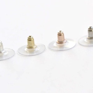 Pode incluir: Quatro fechos de brincos em diferentes acabamentos metálicos: prata, ouro, ouro rosa e prata. Cada fecho tem uma base de disco transparente em forma oval. Os pinos dos brincos são visíveis, mostrando a variedade de tons metálicos.