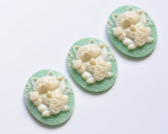May include: Three oval-shaped, green and white ceramic cabochons with a raised design of a white lucky cat holding a gold coin and a paw raised.