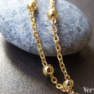 May include: Close-up of a gold-colored necklace with a delicate chain and spherical beads. The necklace is draped over a gray stone, with the VeryCharms logo visible in the bottom right corner.