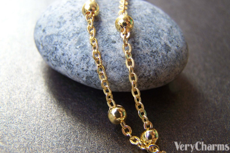 May include: Close-up of a gold-colored necklace with a delicate chain and spherical beads. The necklace is draped over a gray stone, with the VeryCharms logo visible in the bottom right corner.