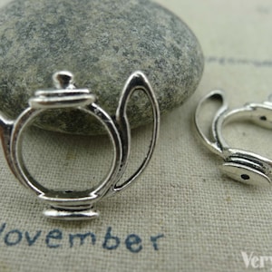 May include: Two silver teapot charms, one with a detailed design and the other a simple outline. The charms are on a textured fabric background.