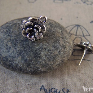 May include: Silver flower charm with a loop for attaching to jewelry or other crafts. The charm is shown on a gray stone.