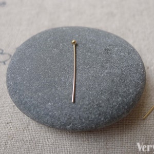 May include: Two gold head pins with round heads. The pins are lying on a gray stone.