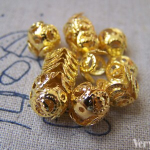 May include: A group of gold-colored metal beads with intricate designs. The beads are round and have a hollow center. They are perfect for jewelry making or other craft projects.