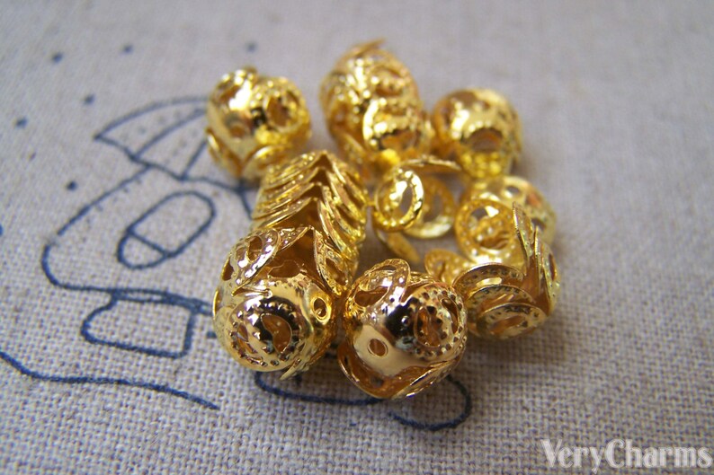 May include: A group of gold-colored metal beads with intricate designs. The beads are round and have a hollow center. They are perfect for jewelry making or other craft projects.