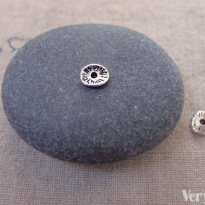 May include: Two small, silver-coloured metal beads with a floral design are displayed on a grey stone. The beads have a central hole and a textured surface, ideal for jewellery making or crafts.