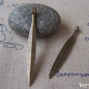 May include: Two antique bronze-coloured metal leaf charms. One charm has a textured, ridged surface, while the other has a striped design. The charms are displayed on a light-coloured fabric with blue drawings.