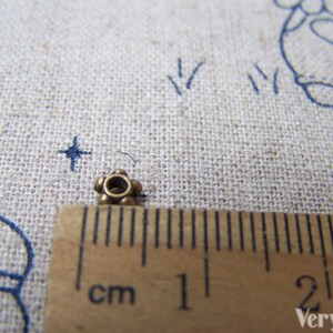 May include: A close-up of a small, gold-colored metal bead with a round hole in the center. The bead is about 1 cm in diameter and has a decorative design of small, raised dots around the edge.