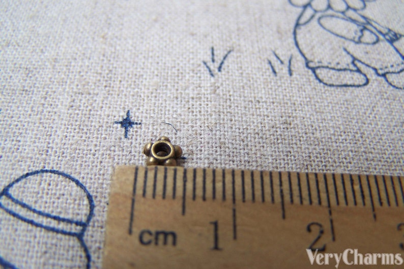 May include: A close-up of a small, gold-colored metal bead with a round hole in the center. The bead is about 1 cm in diameter and has a decorative design of small, raised dots around the edge.