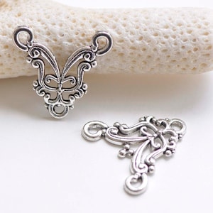 Könnte beinhalten: Zwei silberfarbene Metall-Verbindungsanhänger mit filigranen Designs. Die Anhänger haben eine Schmetterlingsform und eine dreieckige Form.