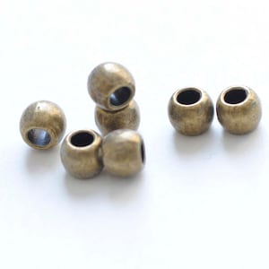 Pode incluir: Um grupo de seis contas de metal de cor dourada antiga. As contas são redondas e têm um furo no centro.