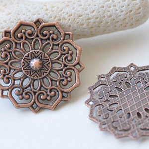 Könnte beinhalten: Zwei kupferfarbene filigranartige Metallverzierungen. Die größere Verzierung hat ein filigraneres Design mit einer kleineren, sternförmigen Mitte. Die kleinere Verzierung hat ein geometrischeres Design.