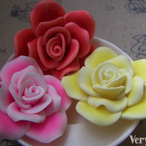 Peut inclure: Trois roses artificielles en fleurs de couleur rose, jaune et rouge. Les roses sont dispos&eacute;es sur une assiette blanche.