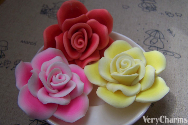 Peut inclure: Trois roses artificielles en fleurs de couleur rose, jaune et rouge. Les roses sont dispos&eacute;es sur une assiette blanche.