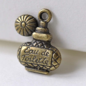 Può includere: Ciondolo in metallo dorato antico a forma di flacone di profumo con un piccolo tappo rotondo. Il flacone ha un motivo a rombi e le parole "Eau de Toilette" incise sul davanti.