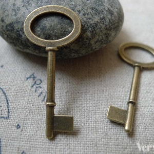 May include: Two antique-style skeleton keys with a brass finish. The keys have a simple design with a round keyhole and a straight shaft. The keys are lying on a textured fabric surface.