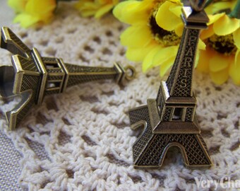 Peut inclure: Deux breloques dorées en forme de Tour Eiffel, l'une avec le mot "PARIS" inscrit sur le côté. Les breloques sont sur une surface en crochet blanc.