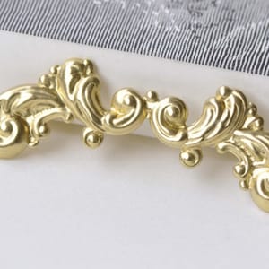 May include: Gold-toned metal decorative trim with a scroll design. The trim has a curved shape and is made of intricate details.