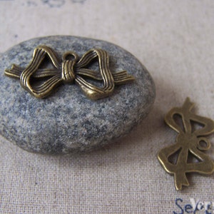 Op de afbeelding: Twee antieke goudkleurige metalen strikcharms. De grotere strikcharm is op een grijze steen. De kleinere strikcharm is op een beige stoffen achtergrond.