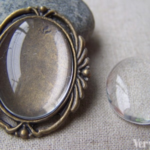 May include: Two glass cabochons, one in a bronze oval frame. The cabochons are clear and round. The frame has a decorative design with small beads.