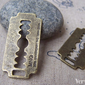 Könnte beinhalten: Zwei antike, goldfarbene Rasierklingen-Anhänger aus Metall. Der größere Anhänger trägt die Gravur "MNQ 20th ANNIVERSARY TAG 1884-2004".