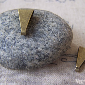 Op de afbeelding: Twee antieke messing hangerbedels, ook wel springringen genoemd, worden getoond op een grijze steen. De bedels zijn driehoekig van vorm met een kleine opening aan de bovenkant.