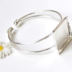 May include: Silver bangle bracelet with a square-shaped glass cabochon setting. The setting is attached to the bracelet on one side.