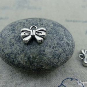 Op de afbeelding: Twee zilveren strikcharms, één op een grijze steen en één op een witte stoffen achtergrond. De strikcharms zijn ongeveer 2,5 cm groot.
