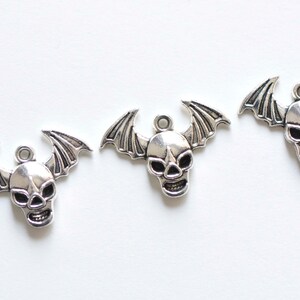 Puede incluir: Tres abalorios de calavera de plata con alas de murciélago. Cada abalorio tiene una calavera detallada con la boca abierta y un par de alas.