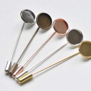 May include: Five round, blank, metal,  cabochon settings attached to silver, gold, and rose gold pins. The settings are silver, gold, bronze, and black.
