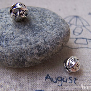 May include: Two silver metal beads with a decorative design. The beads are on a gray rock and a beige fabric surface.