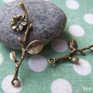 May include: Two antique bronze metal charms in the shape of a branch with leaves and flowers. The charms have a loop at the end for attaching to jewelry or other crafts.