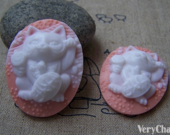 May include: Two white ceramic cat figurines with pink backgrounds. The cats are in a sitting position with their paws raised. The figurines are oval shaped.