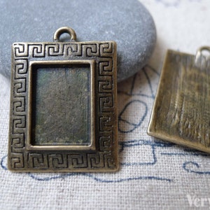 May include: Two antique gold-toned metal picture frame pendants. The frame on the left has a Greek key border and a blank space for a photo or image. The frame on the right has a textured surface.