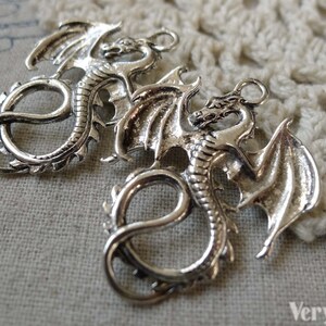 May include: Two silver dragon charms with detailed wings and curled tails. The charms have a loop at the top for attaching to jewellery or other crafts.