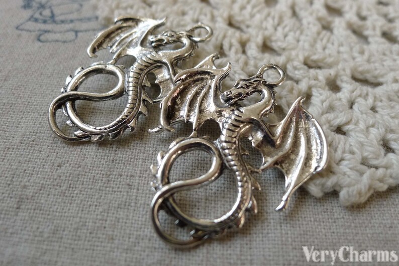 May include: Two silver dragon charms with detailed wings and curled tails. The charms have a loop at the top for attaching to jewellery or other crafts.