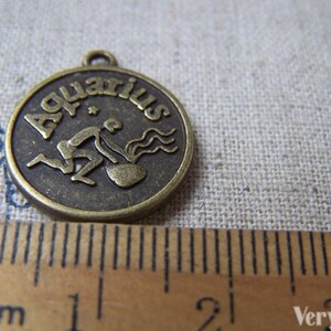 May include: A bronze-colored metal charm with the word "Aquarius" and a stylized image of the water bearer zodiac sign. The charm is approximately 2 cm in diameter.