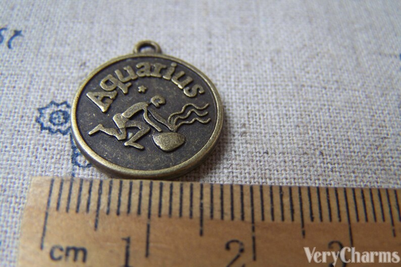 May include: A bronze-colored metal charm with the word "Aquarius" and a stylized image of the water bearer zodiac sign. The charm is approximately 2 cm in diameter.
