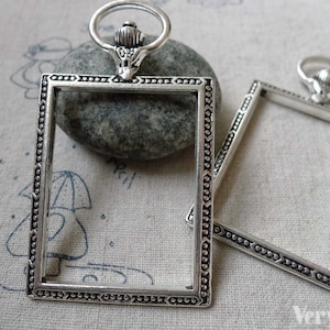 Może przedstawiać: Dwa srebrne metalowe ramki do zdjęć z ozdobnymi krawędziami. Ramki są kwadratowe i mają pierścień u góry do zawieszenia.