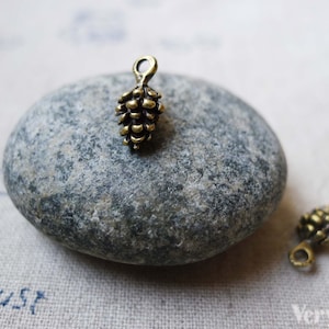 Puede incluir: Dos abalorios de metal de color dorado antiguo en forma de piña. Los abalorios están sobre una piedra gris clara y un fondo de tela blanca.