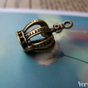May include: A bronze crown charm with a detailed design. The crown is hollow and has a loop at the top for attaching to a necklace or bracelet.