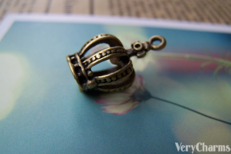 May include: A bronze crown charm with a detailed design. The crown is hollow and has a loop at the top for attaching to a necklace or bracelet.