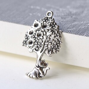 Peut inclure: Breloque en argent représentant un arbre détaillé avec un chat assis sur une branche. L'arbre a un design texturé et orné, et le chat est tourné vers la droite.