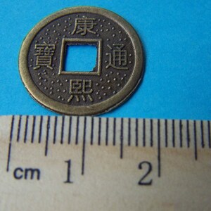 K&ouml;nnte beinhalten: Eine bronzene chinesische M&uuml;nze mit einem quadratischen Loch in der Mitte. Die M&uuml;nze tr&auml;gt chinesische Schriftzeichen, darunter "Wohlstand" und "Gl&uuml;ck". Die M&uuml;nze hat einen Durchmesser von etwa 2,5 cm.