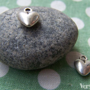 Puede incluir: Dos pequeños colgantes plateados en forma de corazón descansan sobre una piedra gris, uno encima y otro al lado. El fondo es una tela verde con lunares blancos.