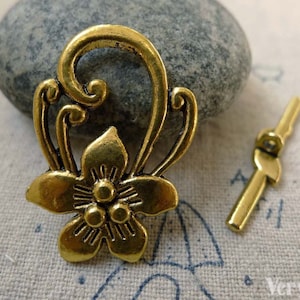 May include: Gold-toned metal flower charm with a decorative swirl design. The charm has a loop at the top for attaching to a necklace or bracelet. A separate gold-toned metal toggle clasp is also included.