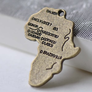 Puede incluir: Un amuleto de metal de color bronce con forma del continente africano. El amuleto tiene una inscripción de varias ciudades y países de África, incluyendo "O CALABAR", "O BRAZZAVILLE" y "O OUAGADOUGOU".