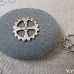 May include: Two silver metal gear charms, one larger than the other, on a gray stone. The larger gear is on the stone, and the smaller gear is on a beige fabric background.